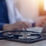 Stethoscope and tablet on desk, doctor using laptop.