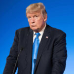 Trump Death Threat – ICE Arrests Illegal Alien! Man in suit with blue background speaking at podium.