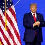 Trump PAUSES ICE Raids – New Directive! Donald Trump in suit near the American flag on stage.