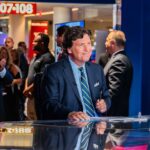 Tucker Carlson Drops BOMBSHELL POLITICAL NEWS People around news desk, Tucker Carlson in suit at the center.