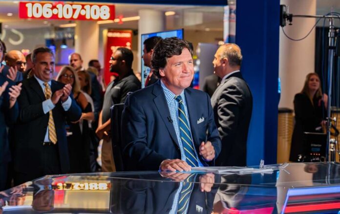 shutterstock_2359655319.jpg featured image People around news desk, Tucker Carlson in suit at the center.
