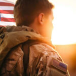 Soldier in camouflage facing American flag at sunset