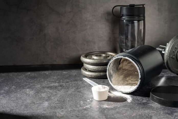 A black protein powder container with a scoop, weights, and a water bottle on a dark surface