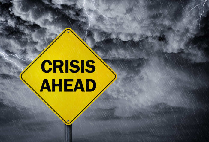 A yellow warning sign that reads 'CRISIS AHEAD' against a stormy sky