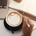 A hand holding a coffee cup with latte art next to a laptop and an open notebook