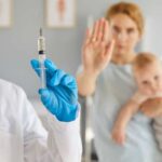 A healthcare professional holding a syringe while a mother with a baby shows a gesture of refusal