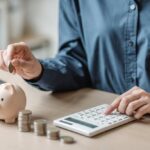 Person placing a coin into a piggy bank while using a calculator