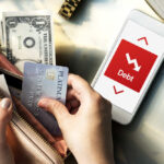 Person holding a credit card and cash while looking at a phone displaying debt information
