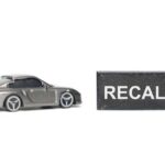 AUTO RECALL: Seat Bolt Disaster A miniature gray sports car next to a black sign that reads 'RECALL'