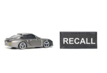 AUTO RECALL: Seat Bolt Disaster A miniature gray sports car next to a black sign that reads 'RECALL'