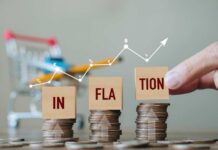 Hand pointing at blocks spelling 'INFLATION' on stacked coins with a shopping cart in the background