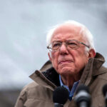 Bernie Sanders wearing glasses speaking outdoors with microphones.