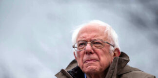 Bernie Sanders wearing glasses speaking outdoors with microphones.