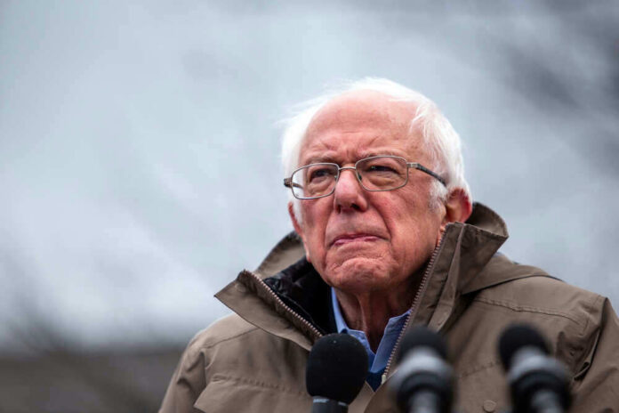 shutterstock_16979420741 Bernie Sanders wearing glasses speaking outdoors with microphones.