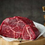 Beef Drops, Chaos Erupts Over Why A raw cut of beef on a wooden board with herbs and a pepper grinder