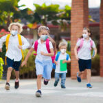 Children with backpacks and masks running outside school.
