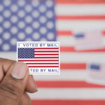 Bombshell Ruling Upends Mail Voting Fight Hand holding a vote-by-mail sticker with U.S. flag