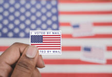 Bombshell Ruling Upends Mail Voting Fight Hand holding a vote-by-mail sticker with U.S. flag