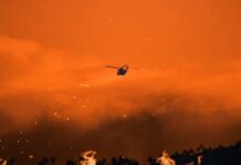 Helicopter fighting massive forest fire with water bucket.