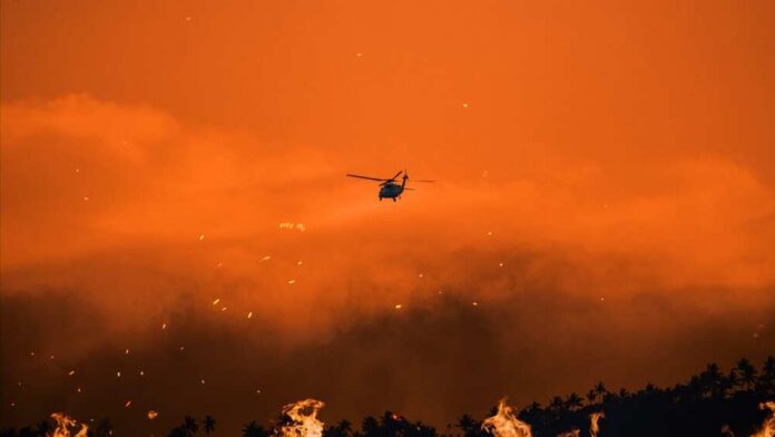 Helicopter fighting massive forest fire with water bucket.