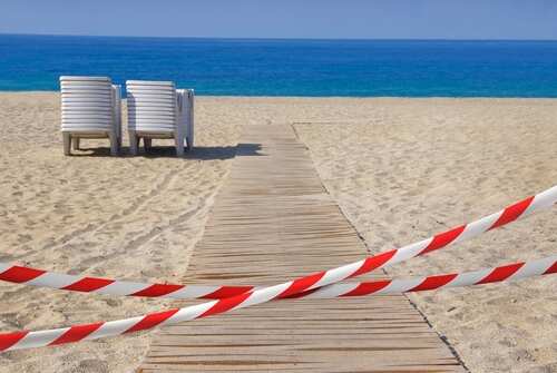 Ribbons,With,Red,And,White,Stripes,Block,The,Entrance,To A wooden pathway leading to the beach with restricted access tape and chairs in the background