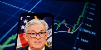 Jerome Powell speaking on a smartphone screen with stock chart in background