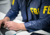 More FBI Turmoil Revealed This Week Person in FBI jacket working on laptop.